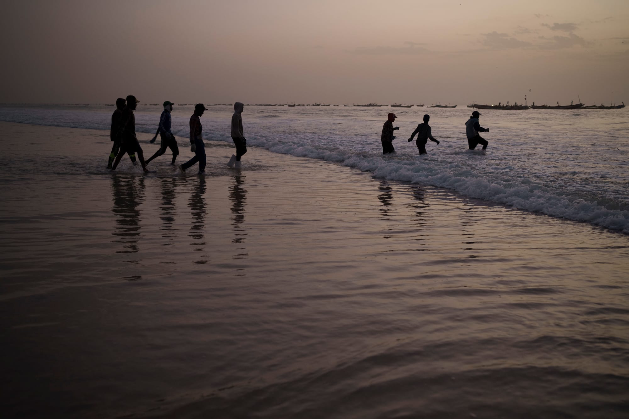 A group of men step into the surf, with fishing boats visible in the distance.