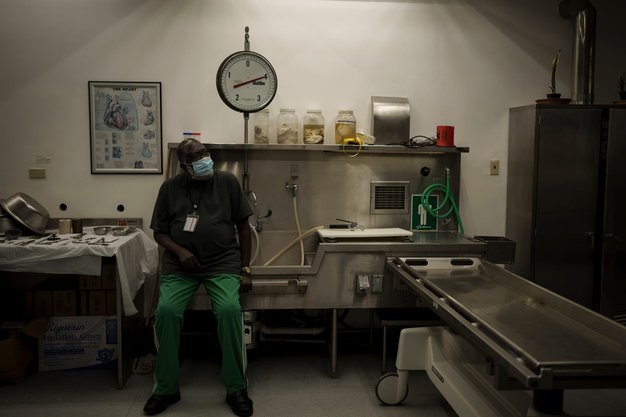 A masked man in scrubs sits in a doctor's office on the edge of an industrial sink.