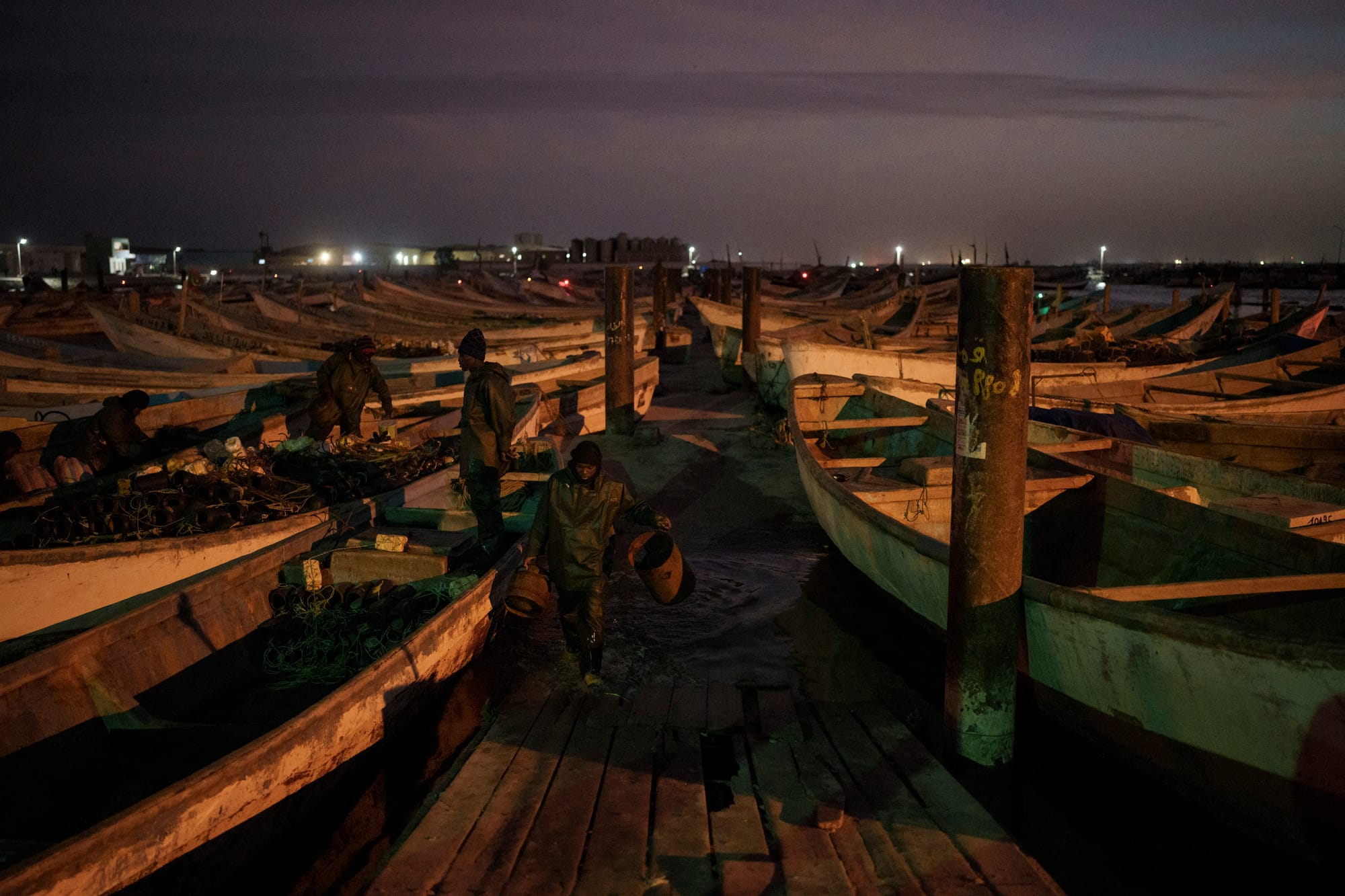 A dock surrounded by fishing boats at dawn. Four fishermen are working and in and around the boats, wearing green jackets.